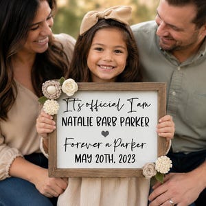 May include: A rectangular wooden sign with a white background featuring black text that reads: It's official I am NATALIE BARB PARKER, a heart symbol, Forever a Parker, and MAY 20TH, 2023. The sign is decorated with small beige and white fabric flowers.