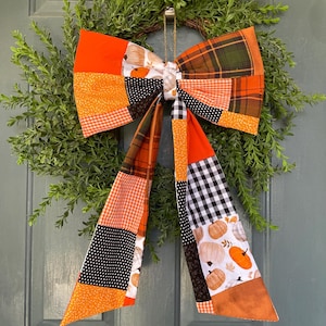 May include: A large decorative bow made from patchwork fabric featuring various patterns including orange solids, black and white gingham, brown plaid, and a white print with orange pumpkins. The bow has two long hanging tails and a central knot.