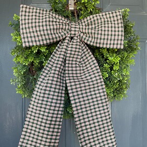 May include: A decorative wreath with a lush green base and a large plaid bow. The bow features a pattern of dark green, red, and white checks. The wreath is hanging on a gray door, creating a welcoming aesthetic.
