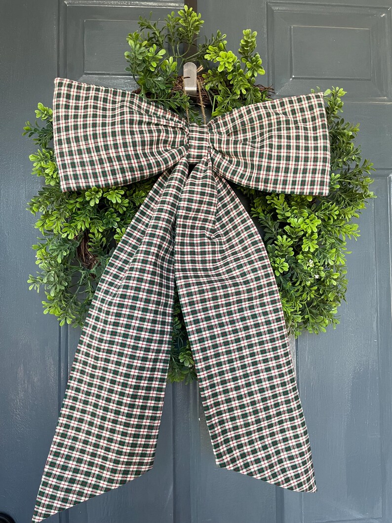 May include: A decorative wreath with a lush green base and a large plaid bow. The bow features a pattern of dark green, red, and white checks. The wreath is hanging on a gray door, creating a welcoming aesthetic.