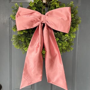 May include: A decorative wreath featuring a lush green base adorned with a large, dusty rose-colored bow. The bow's fabric has a subtle sheen, adding a touch of elegance. The wreath is hanging on a gray door, creating a welcoming aesthetic.