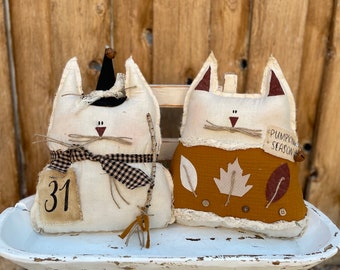 May include: Two stuffed fabric cat dolls. The left cat is cream-colored with a black witch hat, a black and tan checkered scarf, and a tag labeled 31. The right cat is mustard yellow with leaf appliques, lace trim, and a tag reading PUMPKIN SEASON.