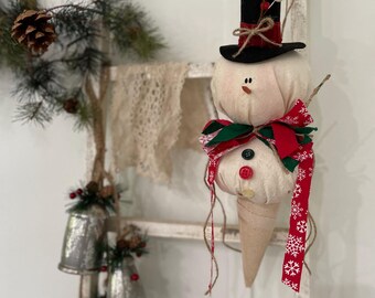 May include: A snowman-shaped Christmas ornament with a black top hat, red and green scarf, and a cone-shaped base. The snowman has button eyes and a carrot nose. The ornament is hanging on a white ladder with other holiday decorations.