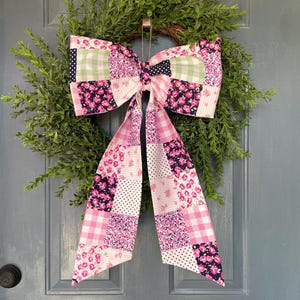 May include: A decorative wreath featuring a large bow made of patchwork fabric in pink, white, and navy blue floral, gingham, and polka dot patterns. The bow is attached to a green, leafy wreath, creating a welcoming home decor piece.