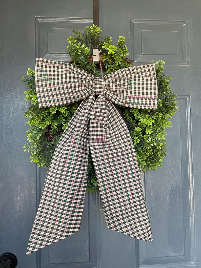 May include: A decorative wreath with a large plaid bow. The bow features a green, red, and white plaid pattern. The wreath is made of green foliage and is hanging on a blue door.