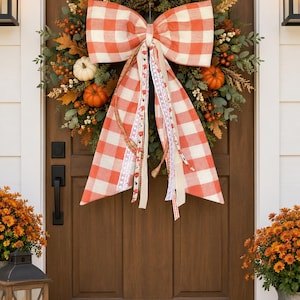 May include: A decorative autumn wreath featuring green foliage, orange berries, and small white and orange pumpkins. A large, prominent bow made of orange and white buffalo check fabric hangs from the center, with long, trailing ribbons in various patterns.