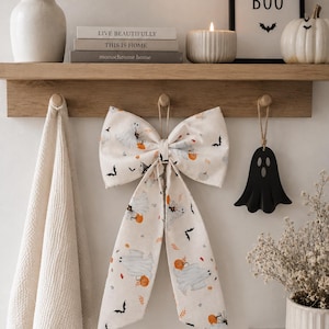 May include: A large fabric bow featuring a repeating pattern of white ghosts, orange pumpkins, black bats, and spiderwebs on a cream background. The bow has two long, hanging tails and is suspended from a wooden hook on a wall-mounted rack.