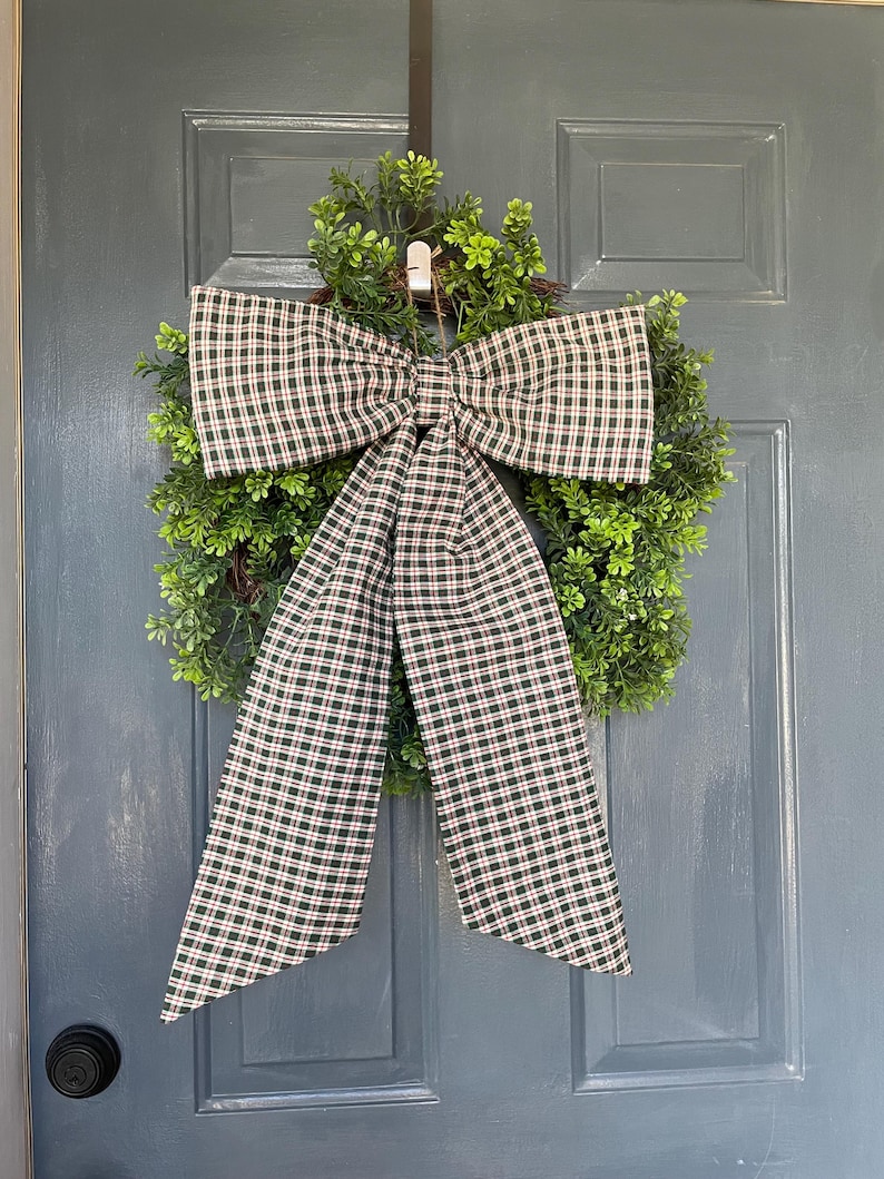 May include: A green wreath with a large plaid bow, hanging on a blue door. The bow features a pattern of black, white, and red squares. The wreath is made of small green leaves.
