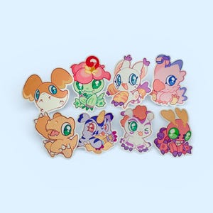 May include: A collection of eight colorful cartoon character pins. Each pin features a unique creature design with vibrant colors, including orange, green, pink, and purple. The pins are arranged on a light blue background, showcasing their detailed artwork.