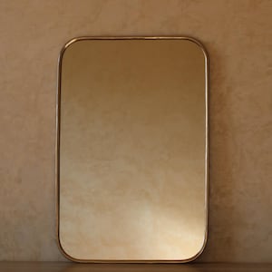 May include: A rectangular mirror with rounded corners and a thin, gold-colored frame. The mirror's surface reflects a warm, golden hue, and the background is a textured, neutral-toned wall.
