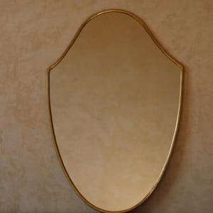 Puede incluir: Un espejo con marco dorado de forma única, similar a un escudo. La superficie reflectante está rodeada por un borde delgado de oro metálico. El diseño es simple pero elegante, adecuado para varios estilos de interiores.