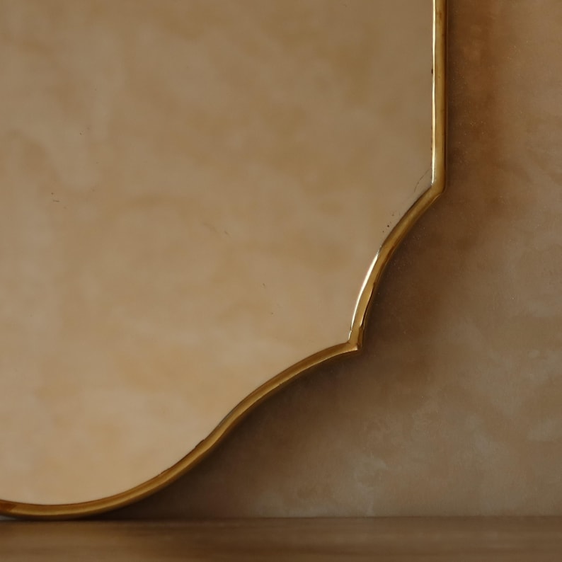 May include: A decorative mirror with a gold-colored, ornate frame. The mirror has a unique, curved shape and a warm, neutral background. The frame's design adds a touch of elegance to the overall aesthetic.