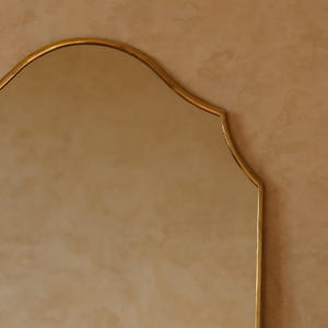 May include: A gold-framed mirror with a scalloped top edge. The mirror's frame has a warm, metallic finish. The background is a neutral, textured wall, providing a subtle contrast to the mirror's reflective surface.