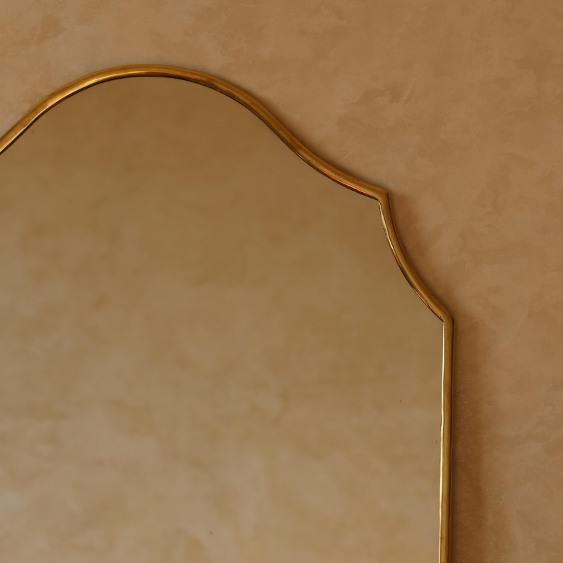 May include: A gold-framed mirror with a scalloped top edge. The mirror's frame has a warm, metallic finish. The background is a neutral, textured wall, providing a subtle contrast to the mirror's reflective surface.