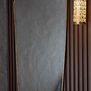 Pode incluir: Um espelho retangular com moldura dourada. O espelho tem uma forma ligeiramente irregular e está montado numa parede de madeira com ripas verticais. À direita do espelho, há uma arandela decorativa com um padrão.