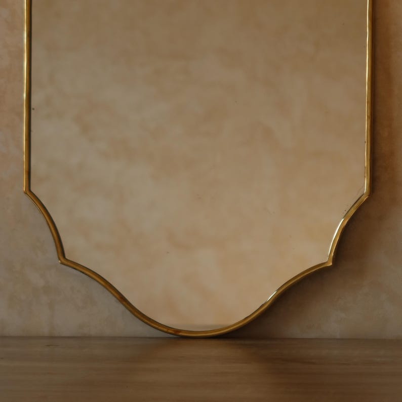 May include: A decorative mirror with a gold-colored frame. The mirror has a unique shield-like shape with a scalloped bottom edge. The frame is thin and elegant, adding a touch of sophistication to the design. The mirror is set against a neutral-toned wall.