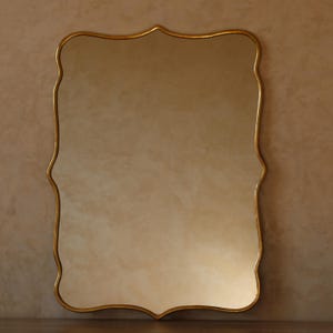 May include: A rectangular mirror with a gold-coloured, ornate frame. The mirror has a unique, scalloped edge design. The glass reflects a soft, warm light, creating a vintage aesthetic. The mirror is a decorative home decor item.
