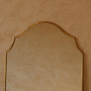 May include: A gold-framed mirror with a distinctive arched top. The mirror's frame has a delicate, ornate design. The background is a warm, neutral tone, providing a subtle contrast to the gold. The mirror is a decorative item.
