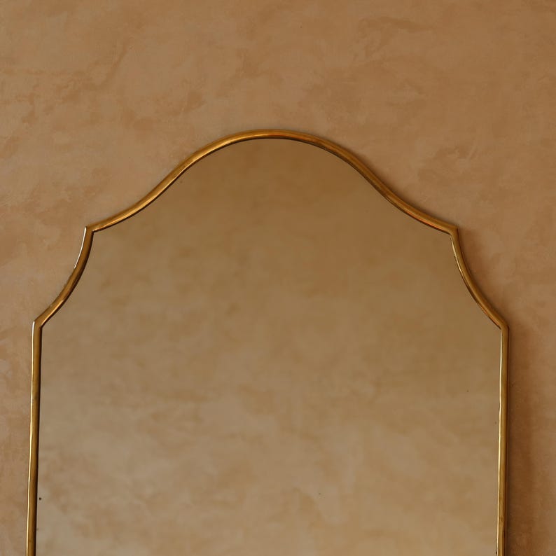 May include: A gold-framed mirror with a distinctive arched top. The mirror's frame has a delicate, ornate design. The background is a warm, neutral tone, providing a subtle contrast to the gold. The mirror is a decorative item.