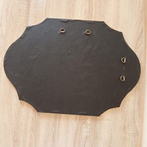 May include: A black, irregularly shaped wall hanging with four metal D-rings for mounting. The back of the piece is visible, showing the dark material and the hardware for hanging. The edges are slightly scalloped.