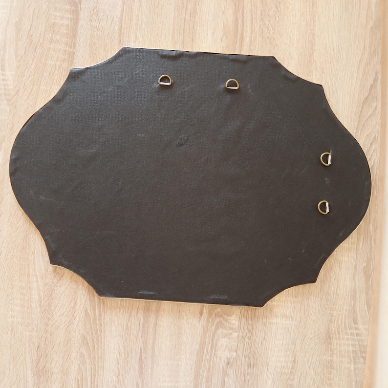 May include: A black, irregularly shaped wall hanging with four metal D-rings for mounting. The back of the piece is visible, showing the dark material and the hardware for hanging. The edges are slightly scalloped.