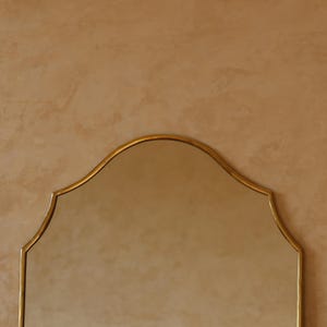 May include: A gold-framed mirror with a distinctive arched top. The mirror's frame has a subtle, elegant design. The background is a warm, neutral color, providing a simple backdrop for the mirror.