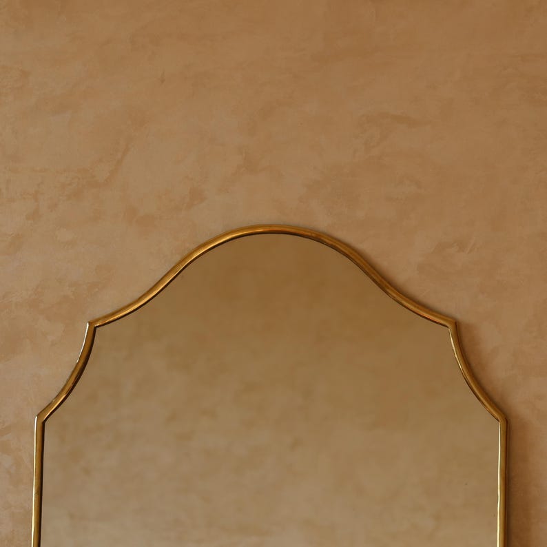May include: A gold-framed mirror with a distinctive arched top. The mirror's frame has a subtle, elegant design. The background is a warm, neutral color, providing a simple backdrop for the mirror.
