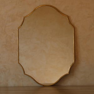 May include: A gold-framed mirror with an ornate, scalloped edge. The mirror has a reflective surface and is set against a neutral-toned wall. The frame has a warm, metallic finish.
