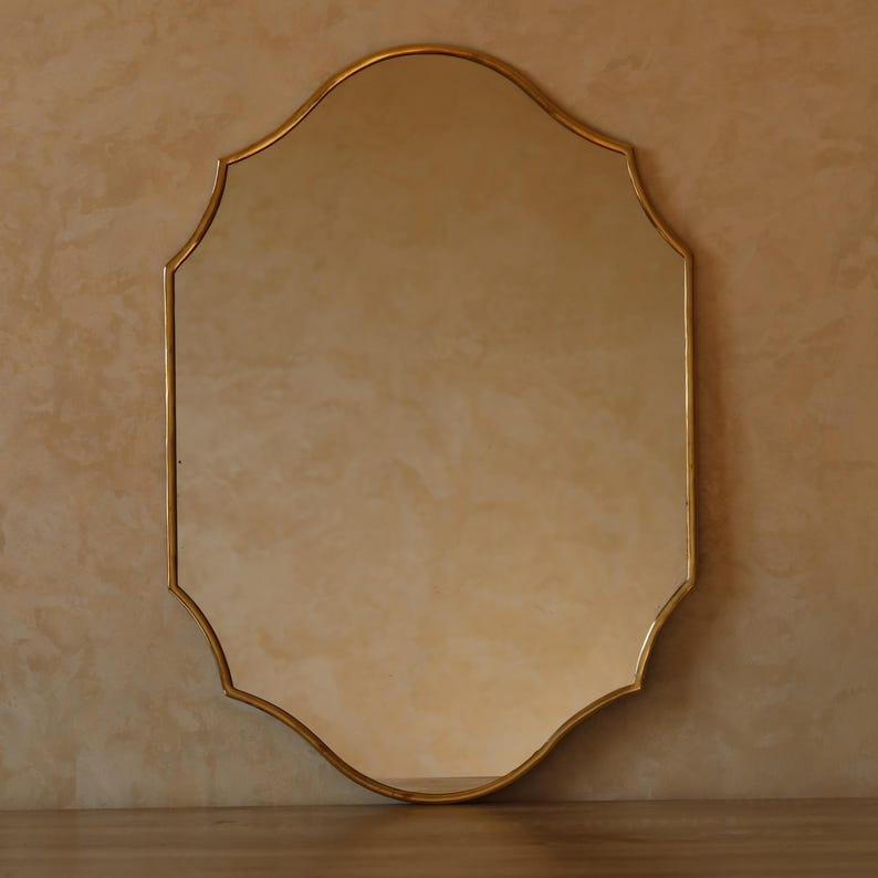 May include: A gold-framed mirror with an ornate, scalloped edge. The mirror has a reflective surface and is set against a neutral-toned wall. The frame has a warm, metallic finish.