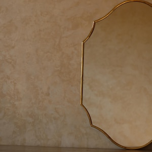 May include: A gold-framed mirror with an ornate, scalloped edge. The mirror's reflective surface is visible, and the frame has a warm, metallic finish. The background is a textured, neutral-toned wall.