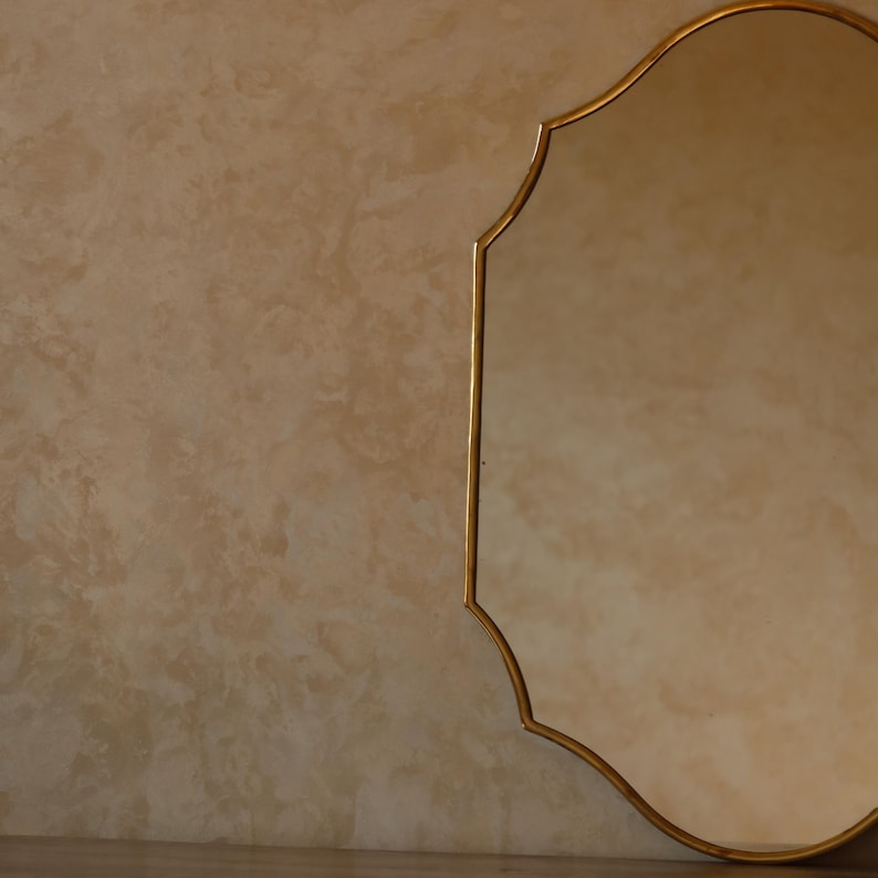 May include: A gold-framed mirror with an ornate, scalloped edge. The mirror's reflective surface is visible, and the frame has a warm, metallic finish. The background is a textured, neutral-toned wall.
