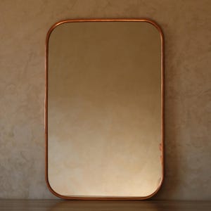 May include: Rectangular mirror with rounded corners, framed in copper. The mirror reflects a soft, warm light, and the copper frame adds a touch of elegance. The mirror is a decorative item for any room.