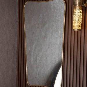 May include: A decorative wall mirror with a gold-colored frame and a unique, curved shape. The mirror is mounted on a dark brown, vertically-slatted wooden wall. A gold-colored sconce with a patterned design is visible on the right.