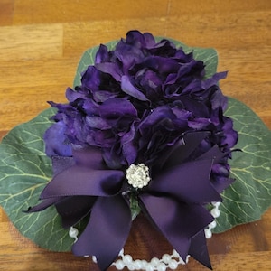 May include: A decorative floral arrangement featuring deep purple hydrangea petals layered over large green leaves. A dark purple satin ribbon bow is centered at the front, accented with a small, round, clear rhinestone embellishment and a string of white faux pearls.