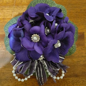 May include: A purple floral arrangement with a green leaf base. The arrangement features multiple purple flower blooms, pearl accents, and a decorative bow with silver and purple ribbons. The arrangement is likely for a special occasion.