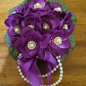 May include: A purple fabric flower corsage featuring multiple layered petals, centered with small white faux pearls surrounded by gold-toned rhinestone settings. The arrangement includes green fabric leaves and a purple ribbon with a dangling pearl strand.