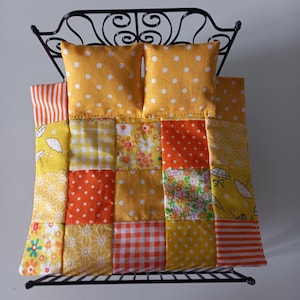 Könnte beinhalten: Miniaturbett mit schwarzem Metallrahmen, zwei gelben Kissen mit weißen Punkten und einer Patchwork-Decke. Die Decke zeigt verschiedene Muster in Gelb, Orange und Weiß, darunter Punkte, Blumenmuster und Streifen. Ein handgefertigter Artikel.