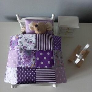 Peut inclure: Un lit miniature blanc avec une courtepointe patchwork dans les tons de violet, blanc et gris. Un animal en peluche avec une tête de souris repose sur un oreiller violet. Une petite table de chevet blanche avec trois tiroirs est à côté du lit. Un jouet en bois est à droite.