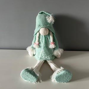 May include: A mint green gnome doll with a knitted hat and dress. The doll has white braids with pink bows, white fluffy trim, and matching feet. The gnome is sitting on a white surface against a gray background.