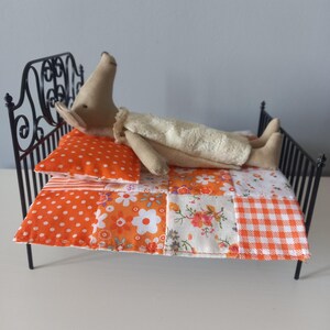 May include: A miniature black metal bed with a doll lying on it. The doll is wearing a cream-colored outfit and resting on a patchwork quilt with orange and white patterns. An orange polka dot pillow and a striped pillow are also on the bed.