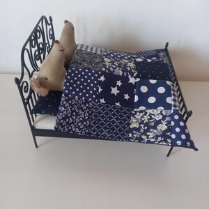 May include: A miniature black metal bed with a patchwork quilt in shades of blue and white. Two beige stuffed animal figures are nestled under the quilt. The quilt features various patterns, including polka dots and stars. A decorative pillow is also visible.