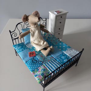 Puede incluir: Una escena de dormitorio en miniatura con una muñeca de ratón de peluche en una cama de metal negro. La cama tiene un edredón de retazos en tonos azules. También hay una pequeña cómoda blanca y gafas. Un libro con cubierta roja está en la cama.