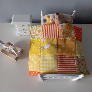Op de afbeelding: Een miniatuur poppenbed met een patchwork quilt in tinten geel, oranje en wit. Een knuffelbeer ligt onder de quilt. Een witte nachtkast met drie lades en een klein houten speelgoed zijn ook aanwezig.