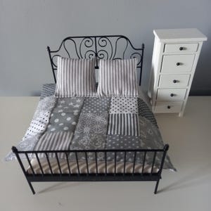 May include: A miniature bed with a black metal frame, a patchwork quilt in grey and white, two striped pillows, and a white chest of drawers. The quilt has various geometric patterns.