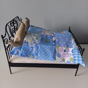 May include: Miniature black metal bed with a white mattress and a patchwork quilt in shades of blue and white. Two small, beige stuffed animals are tucked under the quilt. The quilt features floral, polka dot, and gingham patterns.