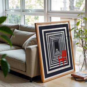 May include: A framed art print featuring an optical illusion of a receding hallway with a red chair. The artwork is in a wooden frame and leans against a window. A beige sofa and greenery are in the background.