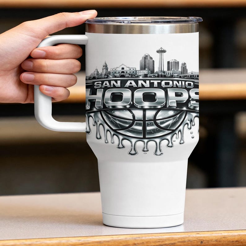 May include: White travel mug with a silver lid and handle. The mug features a black and white graphic of the San Antonio skyline and the text "SAN ANTONIO HOOPS" above a basketball design.