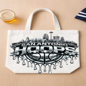 May include: White tote bag featuring a silver and black graphic of the San Antonio skyline and the text "SAN ANTONIO HOOPS" over a basketball design. The bag has white handles and is on a tan surface.