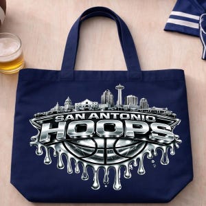 May include: Navy blue tote bag featuring a silver and white graphic of the San Antonio skyline and the text "SAN ANTONIO HOOPS" over a basketball. The bag has two navy blue handles.