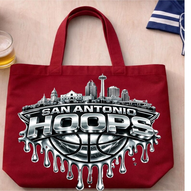 May include: A red tote bag featuring a silver and black graphic of the San Antonio skyline and the text "SAN ANTONIO HOOPS" with a basketball design. The bag has two red handles.